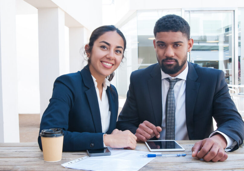 Coworkers working at office desk with tablet, papers and coffee. Business man and woman wearing formal clothes, looking at camera and sitting at desk. Office work concept. Front view.